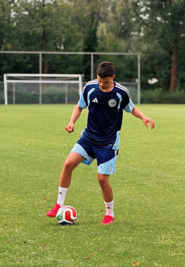 Jeune joueur de football masculin en uniforme bleu contrôle le ballon sur un terrain herbeux avec un but et des arbres en arrière-plan.