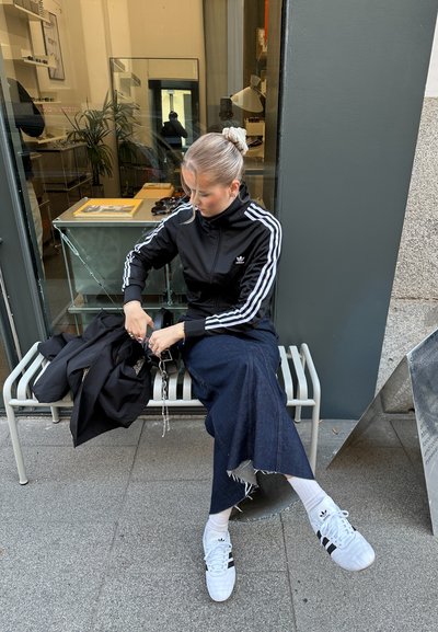 Chaqueta negra con cremallera y rayas blancas, falda de denim y zapatillas blancas. Sentada en un banco, sosteniendo un bolso. Fondo interior minimalista.