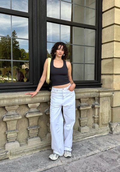 Top sin mangas negro con escote redondo y tachuelas plateadas, pantalones blancos de pierna ancha y zapatillas ligeras, todo contra un fondo de pared de piedra y grandes ventanales.