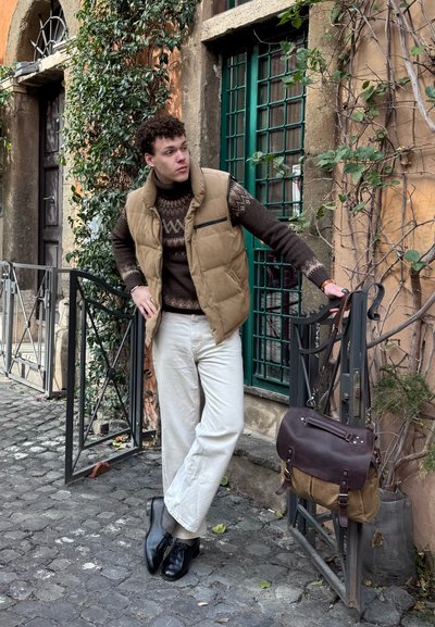 Joven con chaleco beige y pantalones blancos apoyado en una puerta abierta con ventana verde, bolso de cuero marrón colgado en la puerta, camino de adoquines debajo.