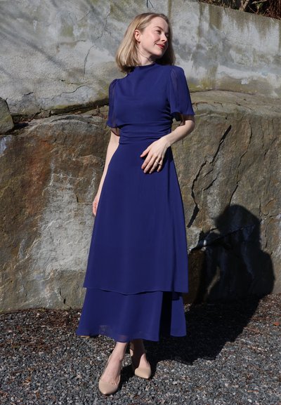 Mujer con pelo corto y rubio, vestida con un vestido largo azul oscuro y tacones beige, está de pie a la luz del sol junto a una pared de piedra, mirando hacia su izquierda.