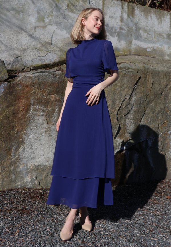 Lühikeste blondide juustega naine, kes kannab pikka tumedat sinist kleiti ja beeže kontsaga kingi, seisab päikese käes kiviseina ääres ja vaatab vasakule.