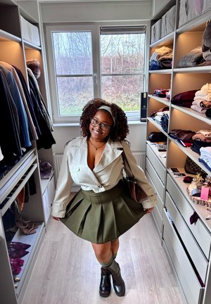 Mujer con cabello rizado y gafas, vestida con una chaqueta beige y una falda oliva, parada en un vestidor con ropa y zapatos organizados.
