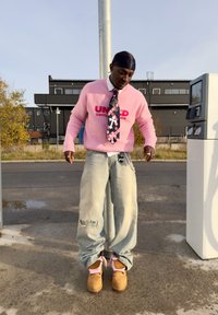 Un homme portant un pull rose, une cravate à motifs, un jean ample déchiré et des bottes brunes se tient près d'une pompe à essence sur une surface en béton.