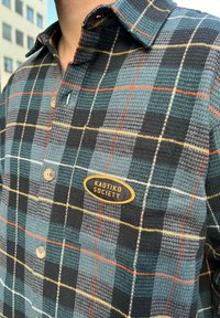 Camisa de botão xadrez em verde-escuro, preto e cinza, com detalhes em laranja e amarelo. Apresenta um rótulo de patch a dizer "KAOTIKO SOCIETY."