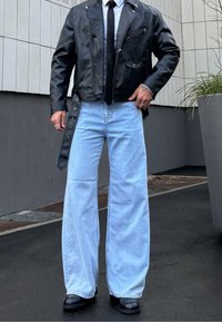 Personne portant une veste en cuir noire, une chemise blanche, une cravate noire, un jean large bleu clair et des bottes noires, debout sur un trottoir sombre en plein air.