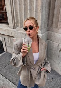 Beige pinstripe blazer met een riem, gedragen over een wit topje. De persoon houdt een groene drank in een doorzichtige beker met een rietje vast. Zwarte zonnebril.
