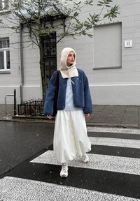 Giacca oversize in pile blu, maglione azzurro chiaro, gonna bianca voluminoso, berretto con cappuccio crema e scarpe da ginnastica bianche su una strisce pedonale. Fiocchi di neve visibili.