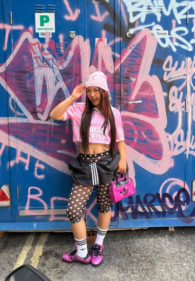 Jersey rosa recortada con el texto "Emirates Better", leggings negros con patrón, falda negra, bolso rosa y zapatillas deportivas rosas. Fondo de grafiti.