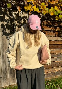 Sweatshirt creme com capuz e bolso tipo canguru, combinado com calças cinzas largas. Boné rosa com o logótipo "LA" e uma bolsa de palha em tom de blush.