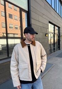 Beige bomullsjakke med en brun kordfløyelskrage, to framlommer, båret over en svart genser, kombinert med lyseblå jeans og en svart caps.