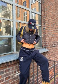 Zwarte cropped denim jack, marineblauwe joggingsbroek met witte NY-logo, en zwarte pet met bloemenontwerp; leunt tegen een bakstenen muur en raam.