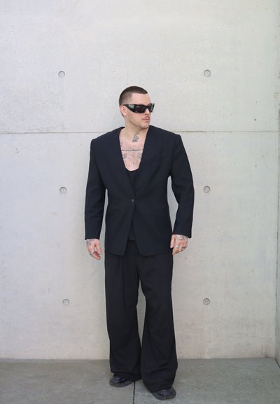 Hombre con traje negro extragrande, gafas de sol negras y zapatos negros está de pie contra una pared de hormigón gris claro mirando hacia un lado.