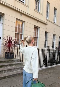 Pull blanc en tricot, jean bleu clair et sac à main vert. Debout sur des marches en pierre devant un bâtiment de couleur claire avec des fenêtres.