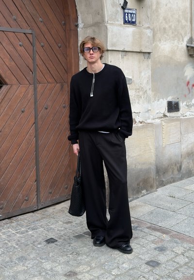 Suéter negro de punto con hombros caídos, pantalones negros holgados y bolsa de malla negra. De pie sobre adoquines cerca de una puerta de madera.