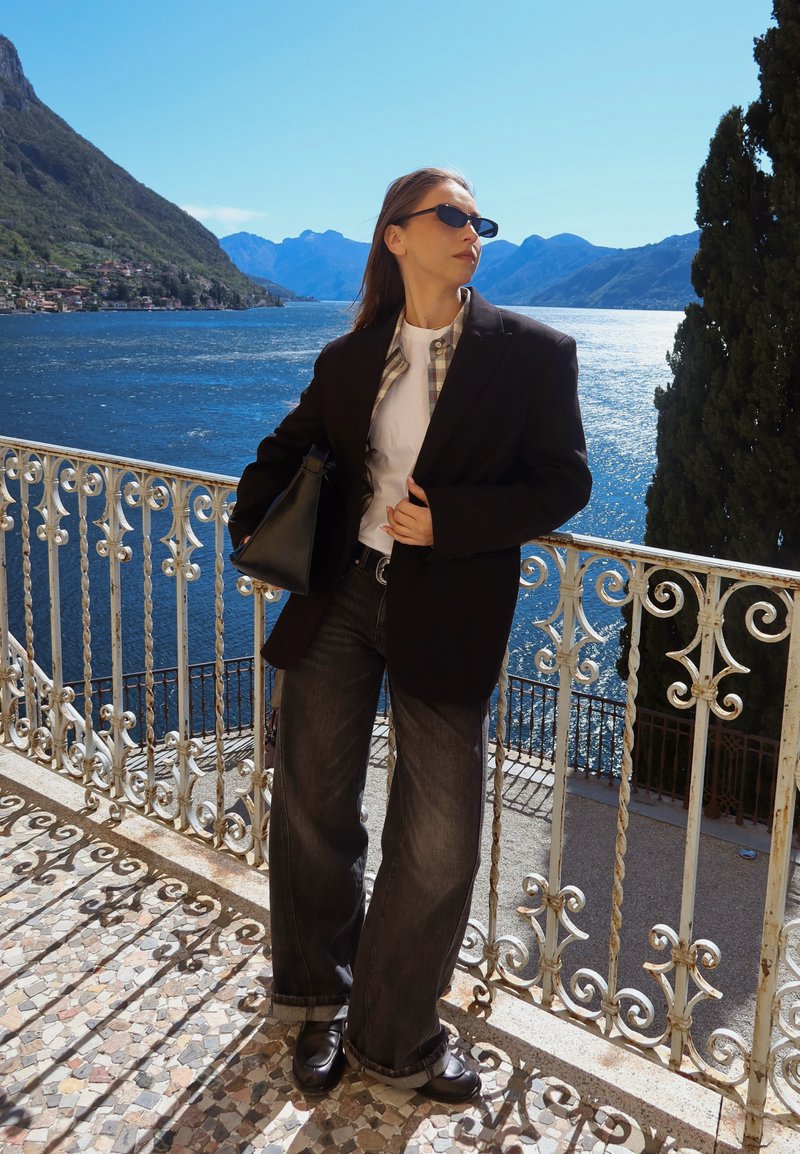 Donna con blazer nero, occhiali da sole, jeans larghi e scarpe nere in piedi su un balcone con vista su un lago blu e montagne.