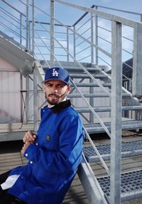 Modrá pracovná bunda s čiernym golierom, zapínaná na gombíky, s vreckom, nosená cez bielu košuľu. Osoba má na sebe modrú čiapku a sedí pri kovových schodoch.