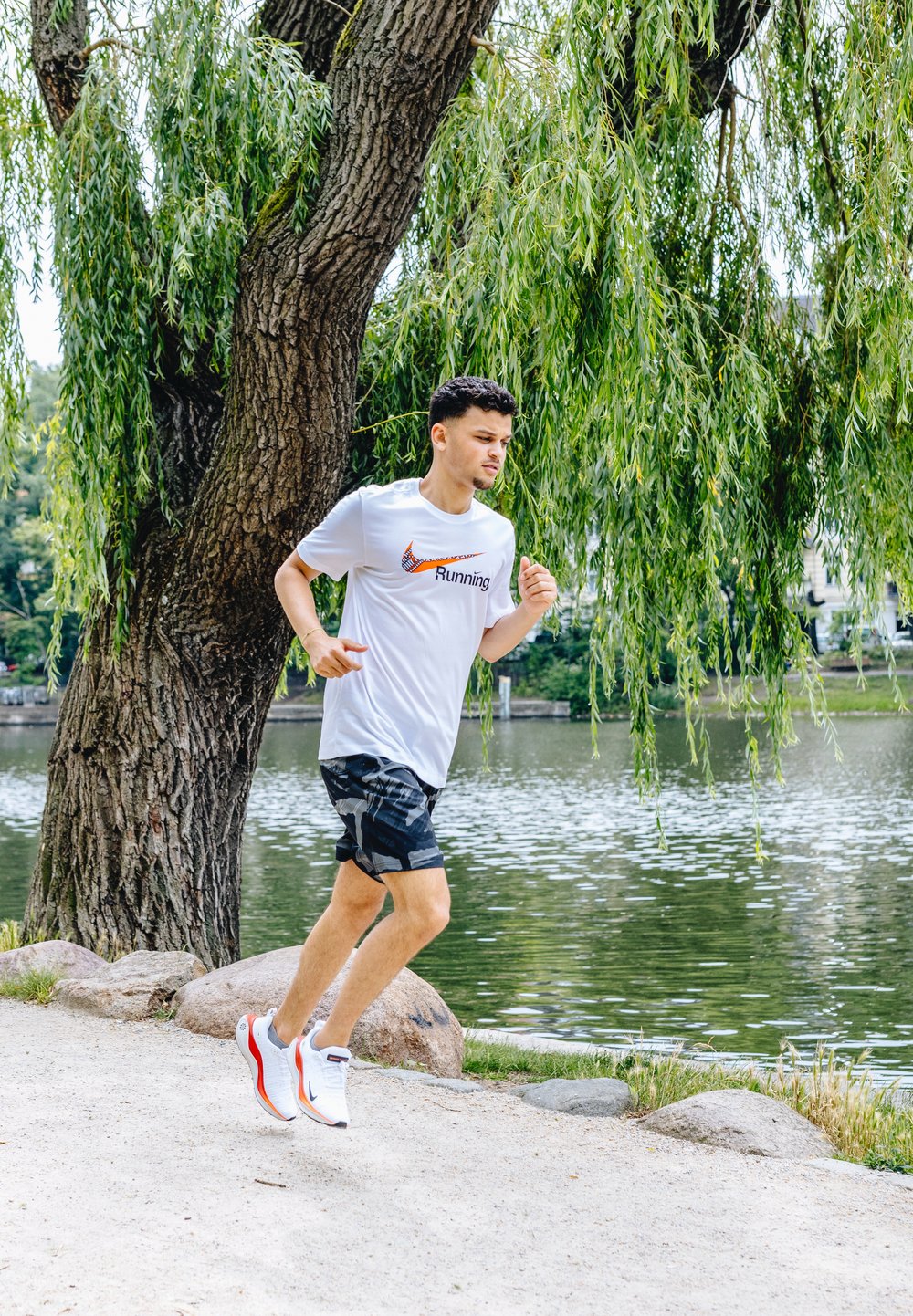 Spordilised jooksujalatsid valges värvuses, millel on oranžid ja sinised aktsendid, koos valge T-särgi ja mustade mustriga lühikestega, asetatud järve äärde.