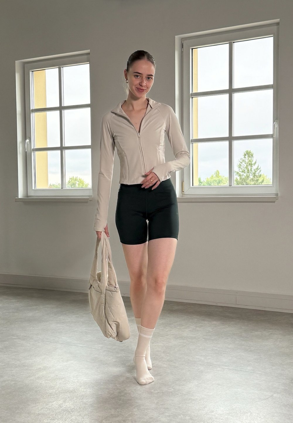Svetlo bež zip-up jakna, črne kratke hlače in bele nogavice do gležnjev. Drži svetlo bež torbo. Notranji prostor z velikimi okni.