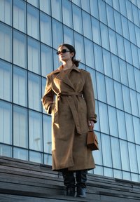 Bruin suède trenchcoat met ceintuur, grote kraag en geruite manchetten; gecombineerd met donkere, nauwsluitende broek en een kleine bruine handtas.