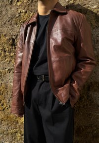 Chaqueta de cuero marrón con cremallera frontal, cuello alzado y bolsillos laterales, combinada con una camisa negra y pantalones, contra una pared texturizada.