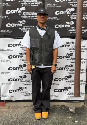 Chaleco negro y gris con bolsillos sobre una camiseta blanca, pantalones negros y botas amarillas. Una gorra negra y un pañuelo completan el atuendo.