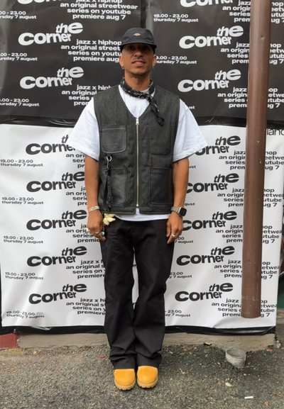 Chaleco negro y gris con bolsillos sobre una camiseta blanca, pantalones negros y botas amarillas. Una gorra negra y un pañuelo completan el atuendo.