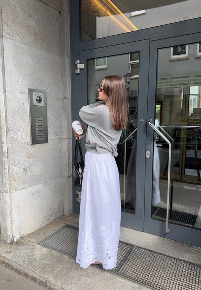 Suéter de punto gris, falda maxi blanca con dobladillo bordado, bolso negro, sosteniendo una taza, de pie al lado de una puerta de cristal y un intercomunicador.