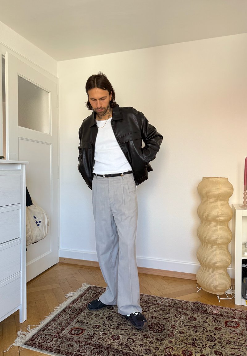 Homme en veste en cuir noire, chemise blanche, pantalon gris et baskets, debout sur un tapis à motifs dans une pièce minimaliste avec une lampe ondulée.