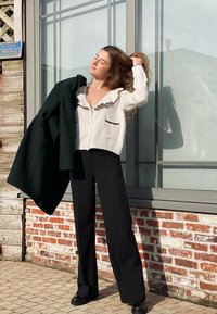 Weißer Cardigan mit Rüschenausschnitt und schwarzem Saum, kombiniert mit schwarzen, hoch geschnittenen Hosen und einem dunkelgrünen Mantel, der über den Arm gelegt ist.