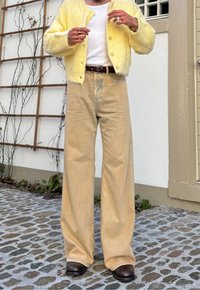 Άτομο με κίτρινη χνουδωτή ζακέτα, λευκό πουκάμισο, φαρδιά μπεζ παντελόνια, σκούρα καφέ παπούτσια, που στέκεται σε πλακόστρωτο πλακ όστρωτο κοντά σε λευκό τοίχο με πέργκολα.