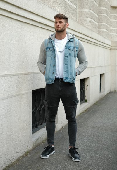 Chaqueta de mezclilla con mangas grises, usada sobre una camiseta blanca, pantalones cargo ajustados negros y zapatillas negras con suelas blancas.