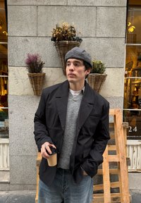 A black blazer worn over a grey sweater, teamed with light blue jeans. Holding a paper coffee cup. Woven planters containing dried flowers in the background.