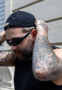 Black cap, black sunglasses, short-sleeved ribbed black top, intricate arm tattoos, visible silver jewellery, light-coloured hair.