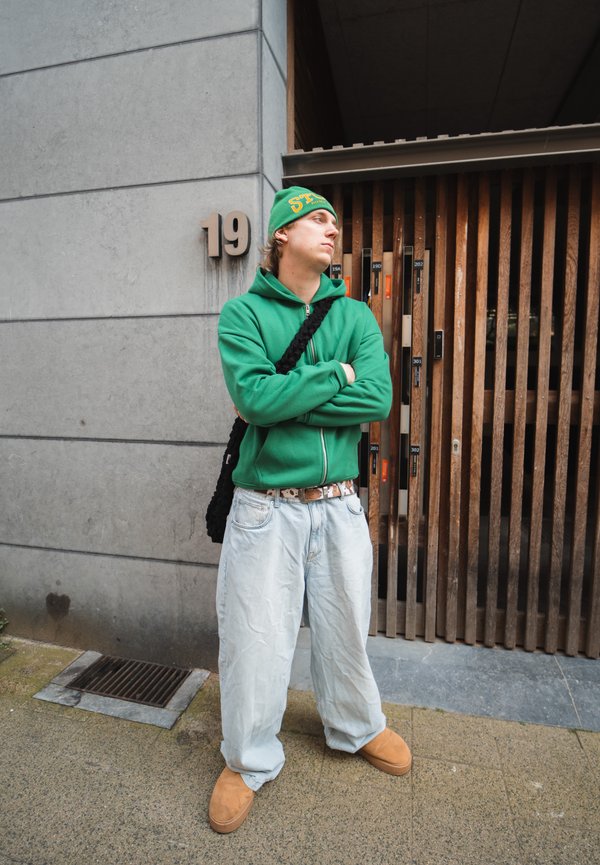 Groene zip-up hoodie met een bijpassende beanie, losse lichtblauwe jeans en bruine instap schoenen voor een houten sleuvendeur met huisnummer 19.