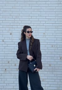 Γυναίκα με μακριά μαλλιά που φοράει γυαλιά ηλίου, σκούρο σακάκι, γκρι πουλόβερ και σκούρο παντελόνι κρατάει μια μαύρη τσάντα χειρός μπροστά σε ανοιχτόχρωμο τοίχο από τούβλα.