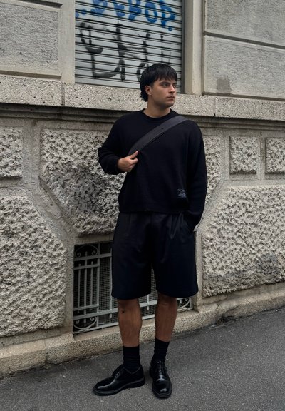 Joven vestido completamente de negro con un bolso al hombro, de pie en una calle frente a una pared de hormigón cubierta de grafitis.
