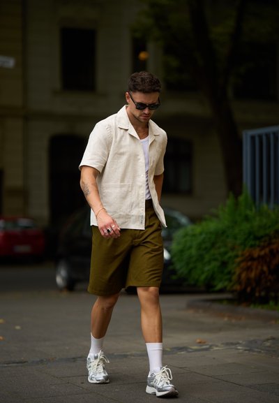 Camisa corta beige claro con un patrón texturizado, combinada con pantalones cortos verde oliva. Calcetines blancos y zapatillas de correr plateadas completan el look.