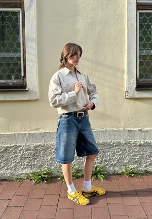 Chaqueta beige clara con cremallera, pantalones cortos de mezclilla, calcetines blancos y zapatillas amarillas con detalles negros. Posicionada contra una pared texturizada beige.