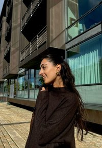Maglione marrone a maglia con maniche lunghe, con polsini a coste. Orecchini dorati, capelli lunghi che cadono, sullo sfondo di un moderno edificio in vetro.