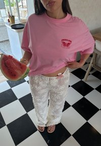 Ανοιχτό ροζ υπερμεγέθη μπλούζα με κόκκινη εκτυπωμένη σχεδίαση πεταλούδας, συνδυασμένη με λευκά υφασμάτινα παντελόνια με φαρδύ πόδι. Κρατώντας μια φέτα καρπούζι.