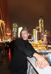 Persona con una giacca nera con colletto e dolcevita, appoggiata a una ringhiera bianca. Anelli alle mani, skyline della città con edifici illuminati sullo sfondo.