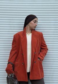 Femme portant un blazer en velours côtelé orange brûlé, un pull crème, un foulard noir et des bijoux en or, tenant un sac à main marron foncé sur un fond gris.