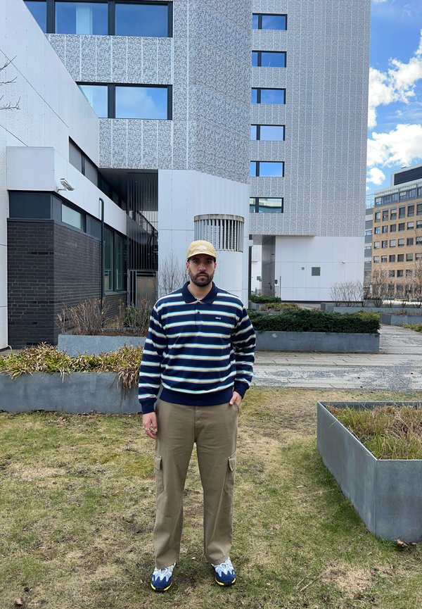 Prugasta svetlo modra in bela jopica, kaki cargo hlače in modro-rumene superge. Stoji pred moderno, sivo-belo stavbo.