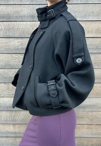 Veste noire texturée avec un col montant, devant boutonné et pattes d'épaule. Caractérisée par des manches larges et des accents de boutons.