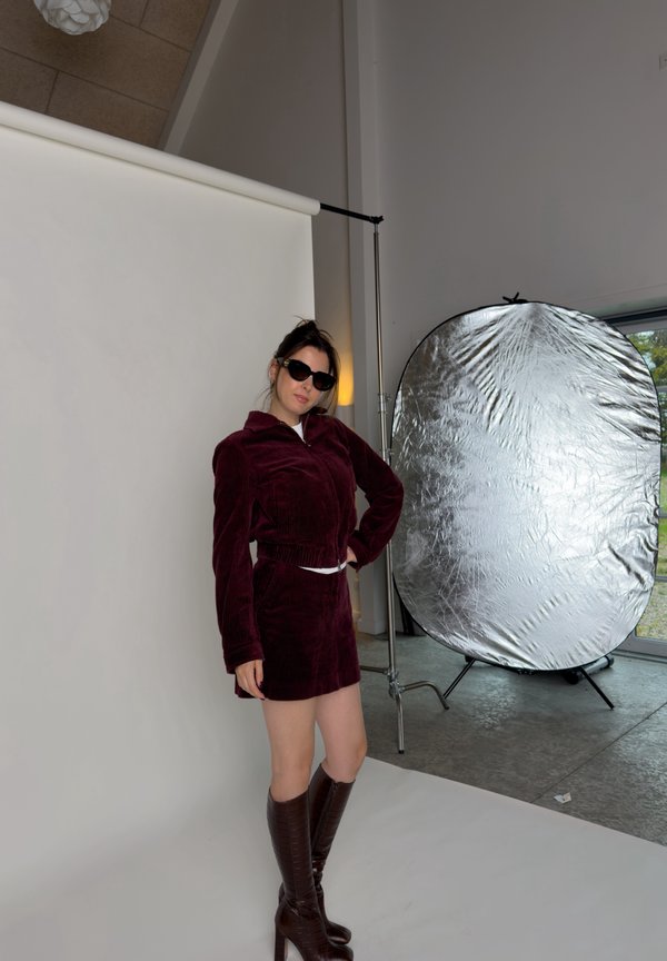 Una mujer con gafas de sol posa con una chaqueta y una falda de color burdeos y botas hasta la rodilla, junto a un gran reflector en un estudio fotográfico.