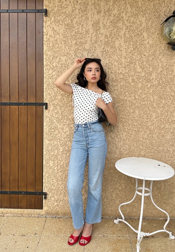 Junge Frau trägt ein gepunktetes Top, hellblaue Jeans und rote Absätze und posiert neben einer strukturierten beigen Wand mit Holztür und rundem weißen Tisch.