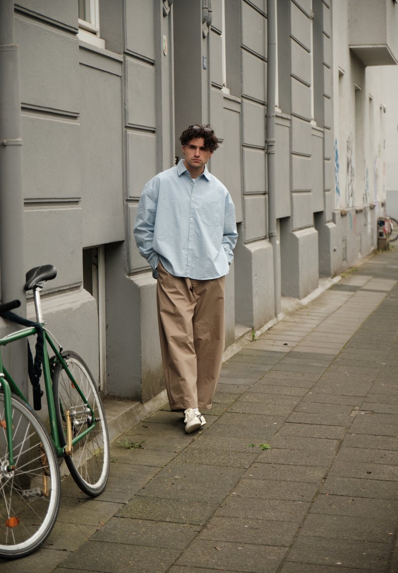 Άνδρας με σγουρά μαλλιά περπατά στο πεζοδρόμιο φορώντας φαρδιά μπλε πουκάμισο, μπεζ παντελόνι, λευκά αθλητικά παπούτσια, δίπλα σε πράσινο ποδήλατο πάνω σε γκρι τοίχο κτιρίου.