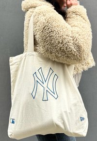 Κρεμ τσάντα καμβάς με μπλε λογότυπο NY, διαθέτει λουριά για ώμους και μια μικρή ετικέτα λογότυπου MLB στη γωνία στο κάτω μέρος. Μαλακή υφή.