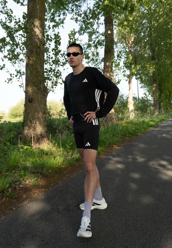 Mann in schwarzer Sportbekleidung und Sonnenbrille steht mit Händen in der Hüfte auf einem von Bäumen gesäumten gepflasterten Weg bei Tageslicht.